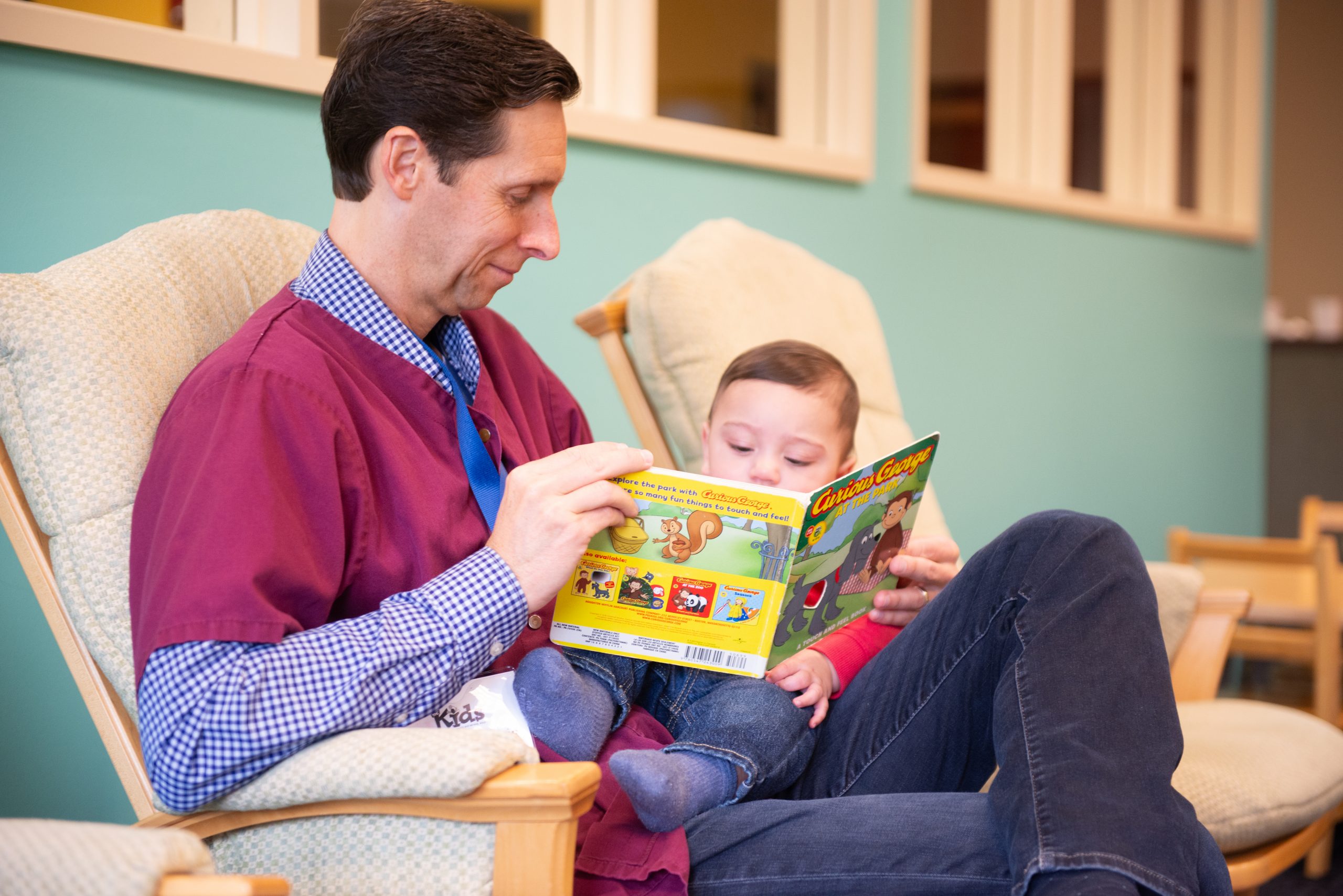 Adult man and young child reading a colorful children's book together in a lounge chair.