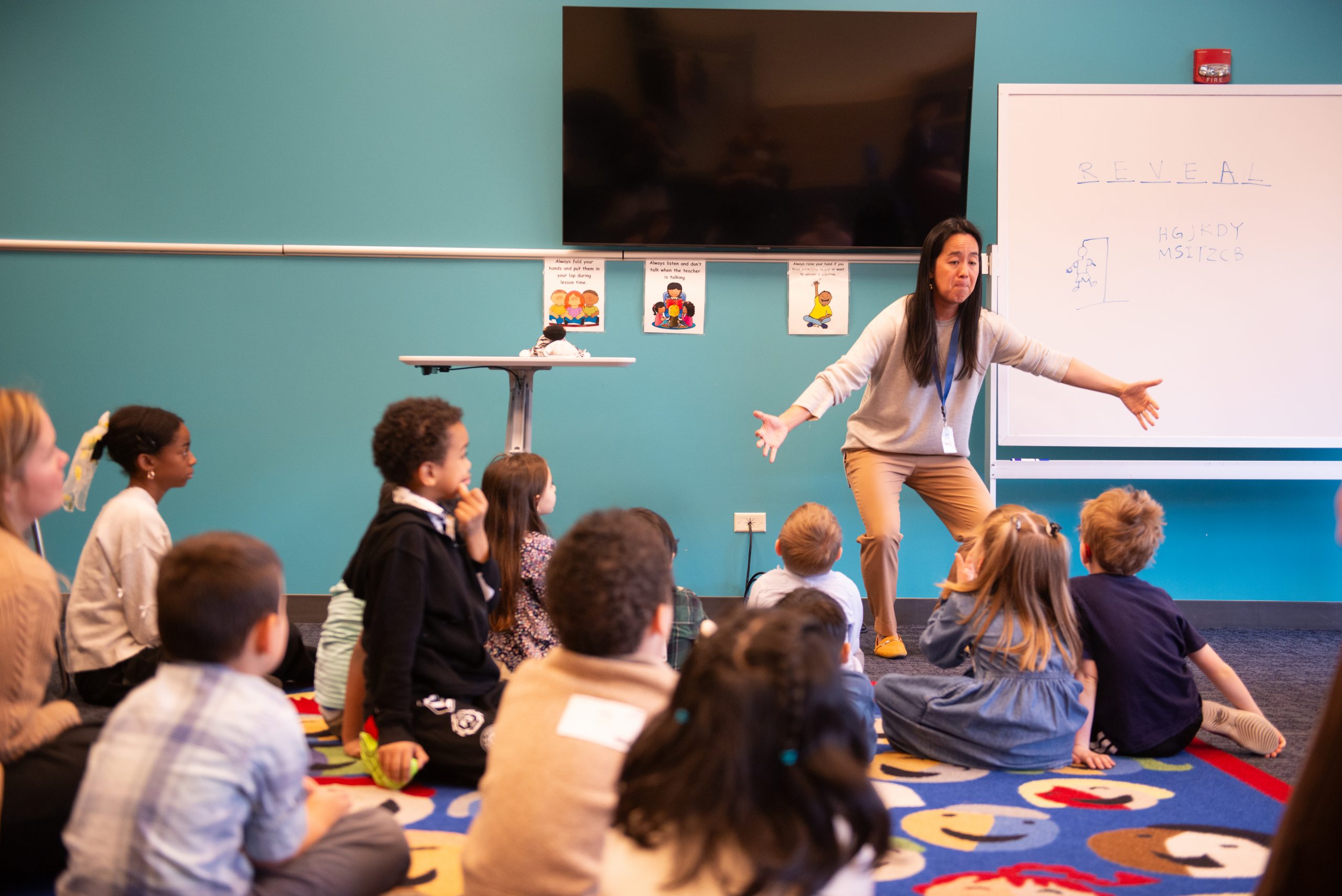 DSC_2251 Teacher at the front with arms outstretched, guiding young students seated on a colorful carpet during a lesson in a teal-wall classroom.