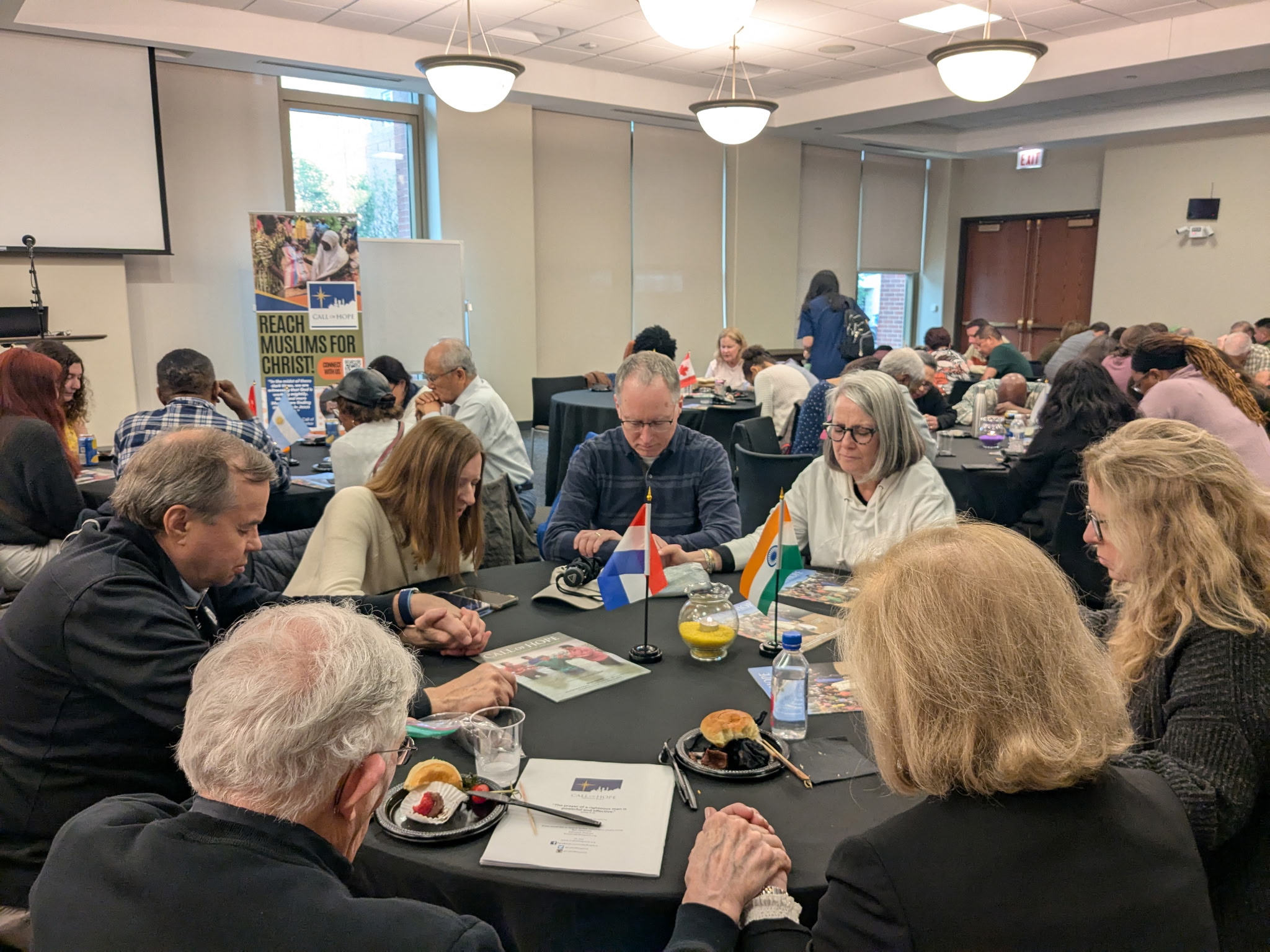6253347859512587209 People seated around tables in a conference room, chatting and taking notes with small flags on the tables in front of them.