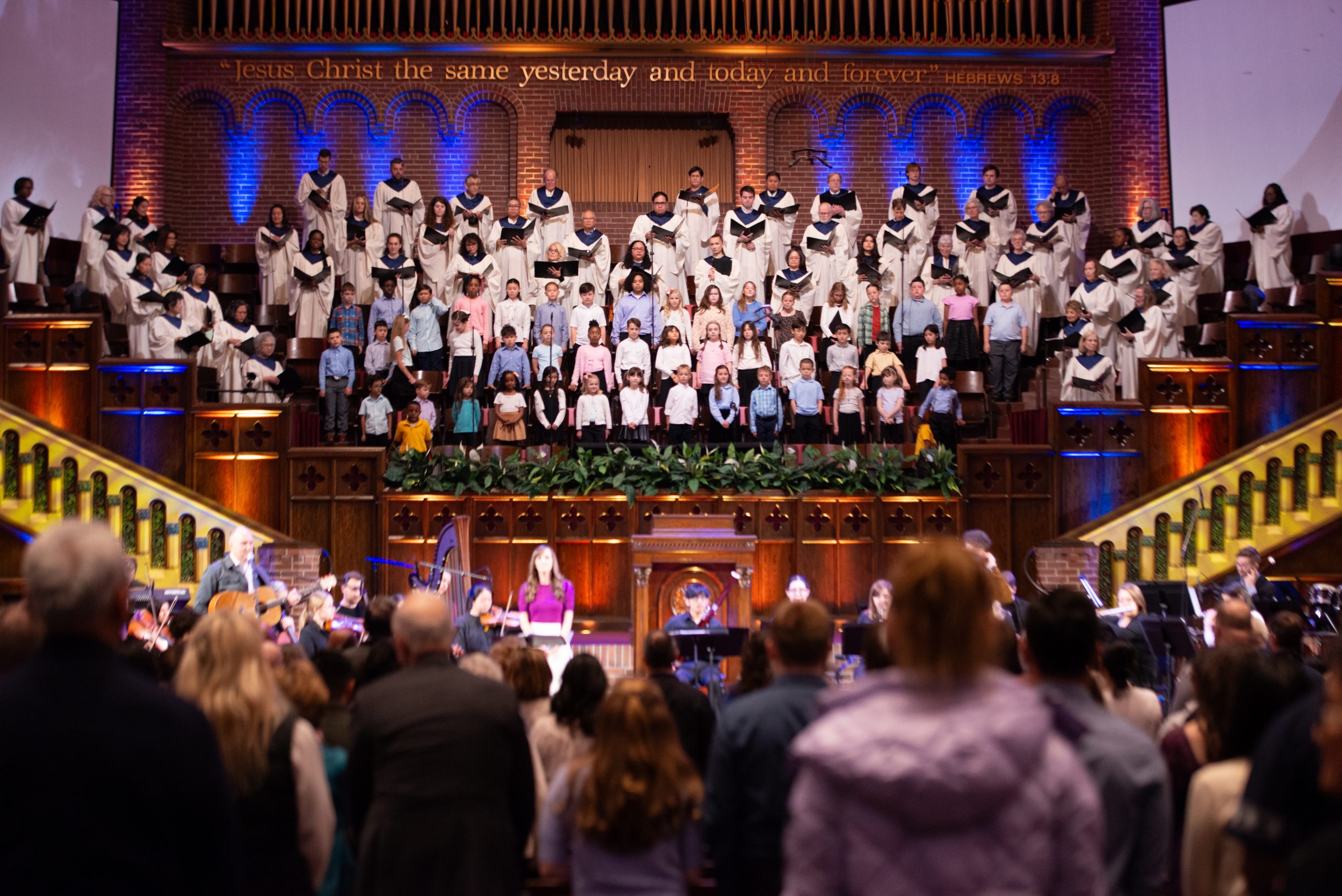 Large choir on a brick church stage, adults in white robes with blue stoles and children in front, performing for an audience.