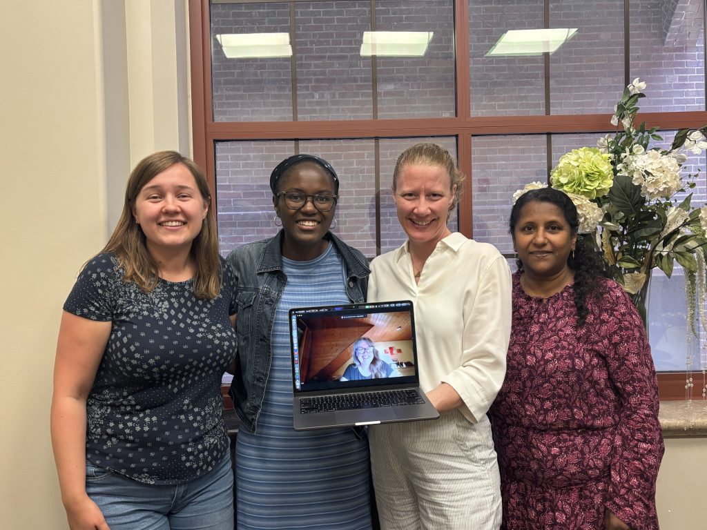 Four smiling women pose indoors, with one holding a laptop showing a video call on the screen behind them.