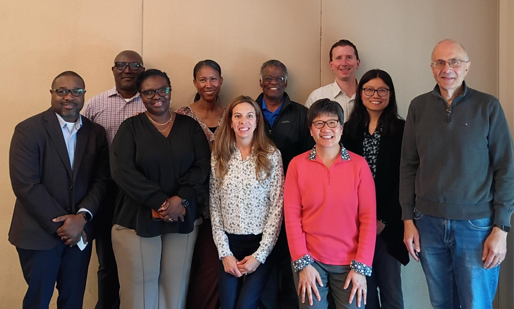 Group of ten professionals posing together for a photo in a neutral indoor setting, smiling at the camera, dressed in business casual attire.