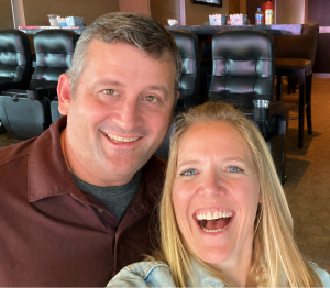 Happy couple taking a selfie together indoors, with rows of black leather chairs and a bar area in the background.