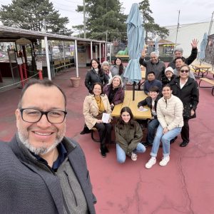 Group of smiling adults and children pose for a selfie in an outdoor area with picnic tables and string lights above.