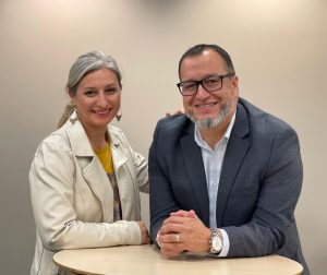 Two smiling adults sit at a round table, a woman with gray hair in a cream jacket beside a man in a dark blazer and glasses.",