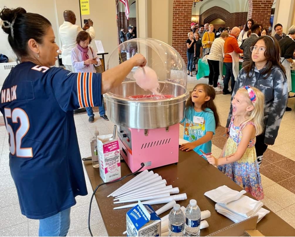 Light & Life Cotton Candy - Moody Fest 2023 Volunteer in a blue sports jersey spins pink cotton candy for smiling children at a community event indoors.