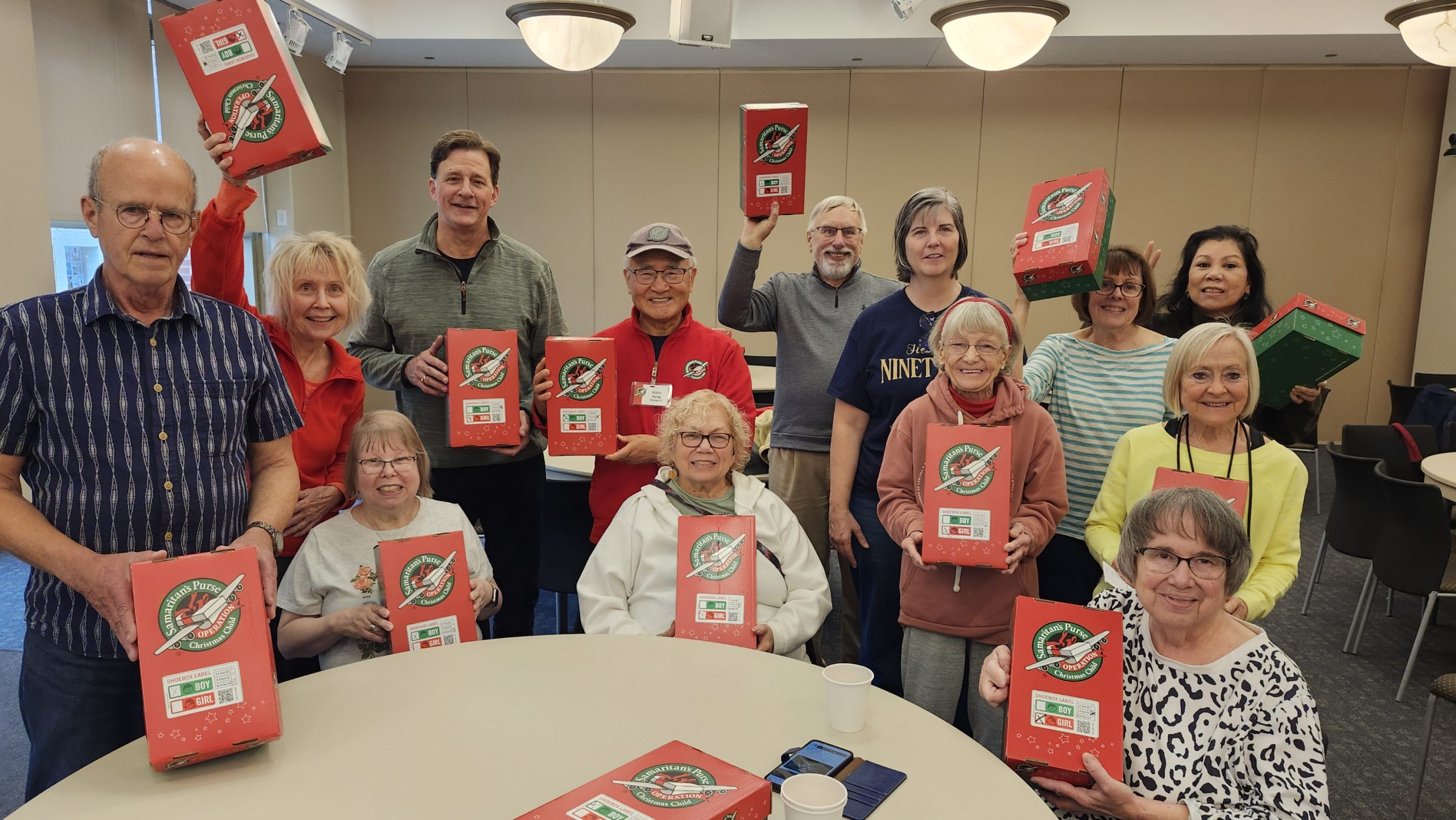 Light & Life Opperation Christmas Child 11-15-2025 Group of smiling volunteers holding red Christmas gift boxes in a community room with tables and chairs visible in the background.