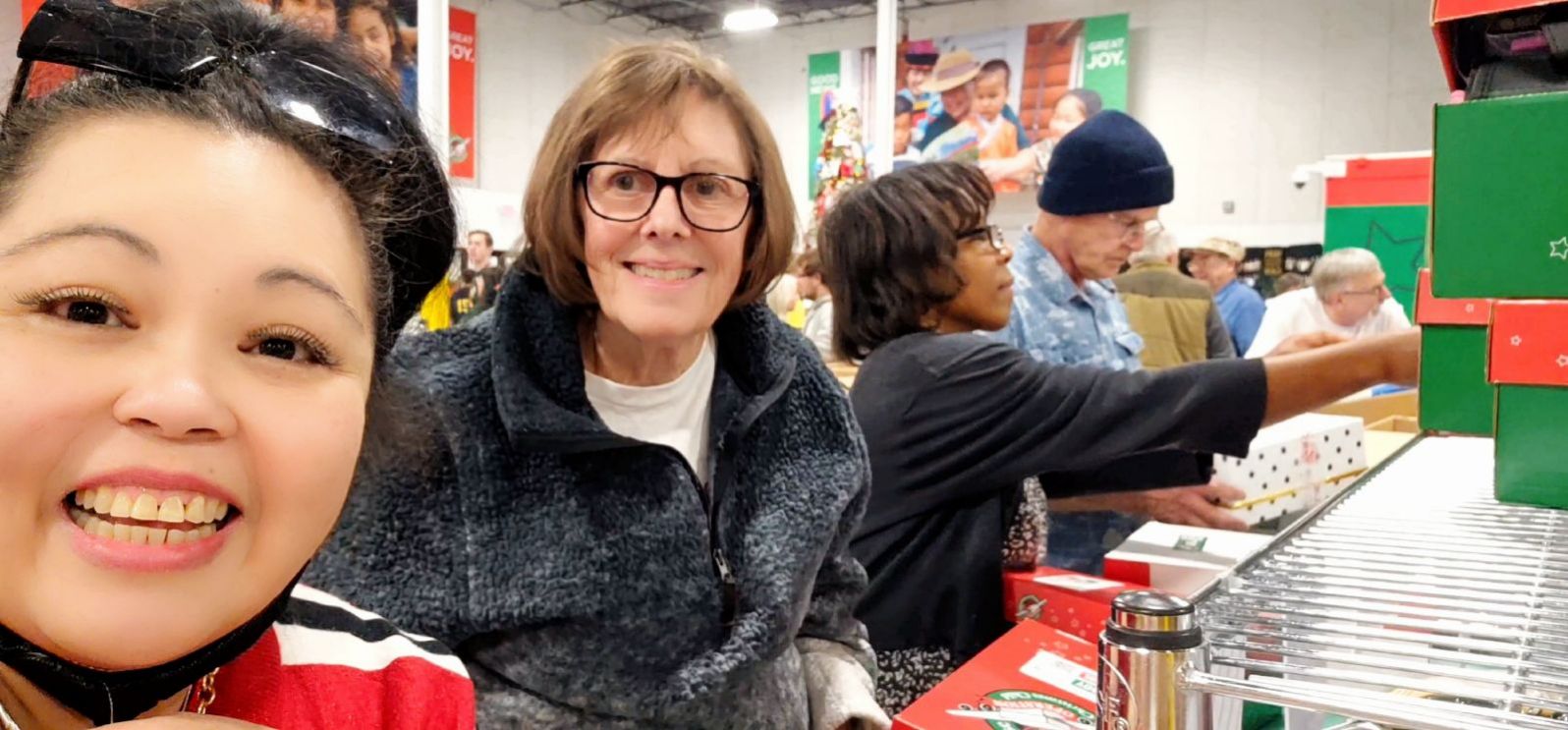 Light & Life - Faith, Cathy, Enosha, & David at Opporation Christmas Child Packing - 12-10-2023 Smiling woman in the foreground taking a selfie with shoppers in a store, where a conveyor of colorful holiday gift boxes is visible in the background.
