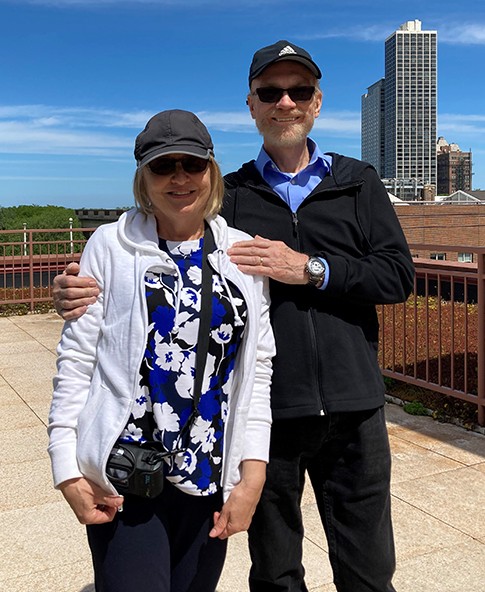 An older couple posing on a sunny rooftop with a city skyline behind them, arms around each other, wearing hats and sunglasses.