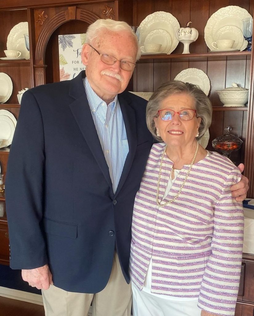 Smiling elderly man in a navy blazer and light shirt stands beside an elderly woman in a striped lavender jacket and pearl necklace, posing arm-in-arm in a dining room with a china cabinet.