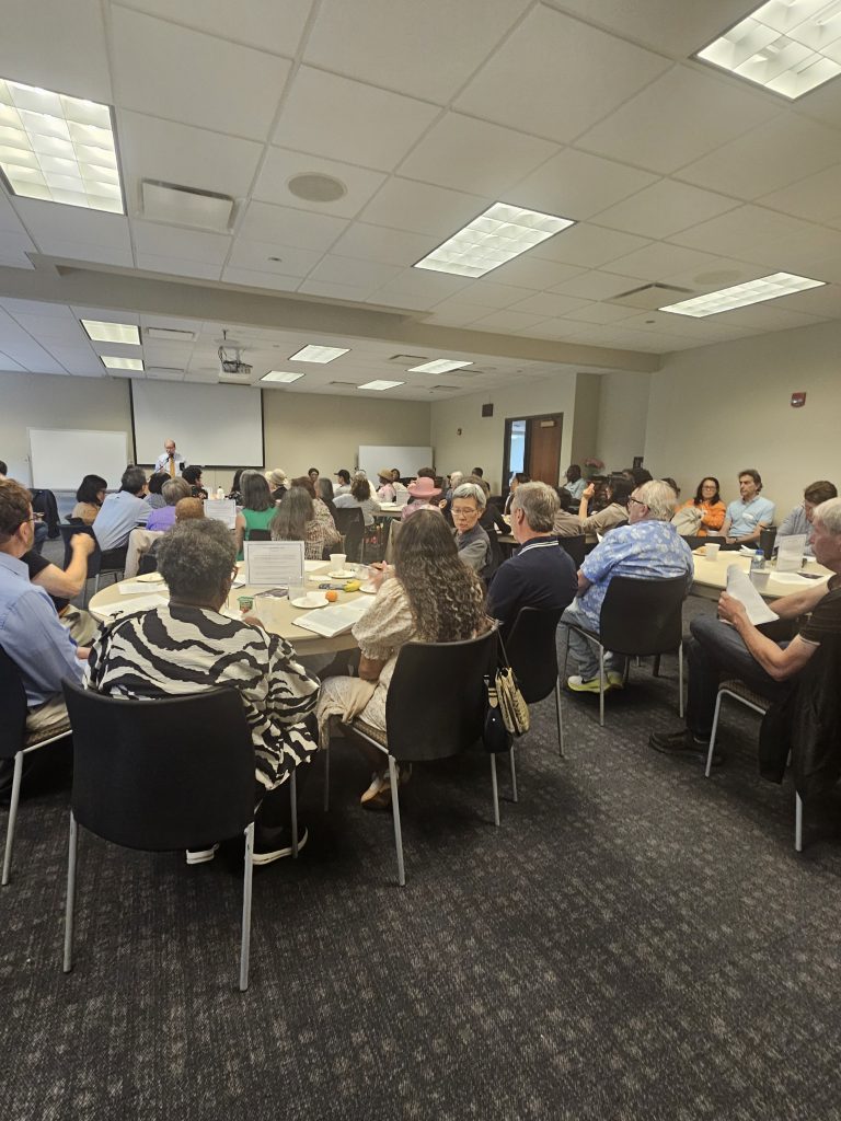 ATM Class3 Large meeting room with round tables and many attendees listening to a speaker at the front, projector screen behind him
