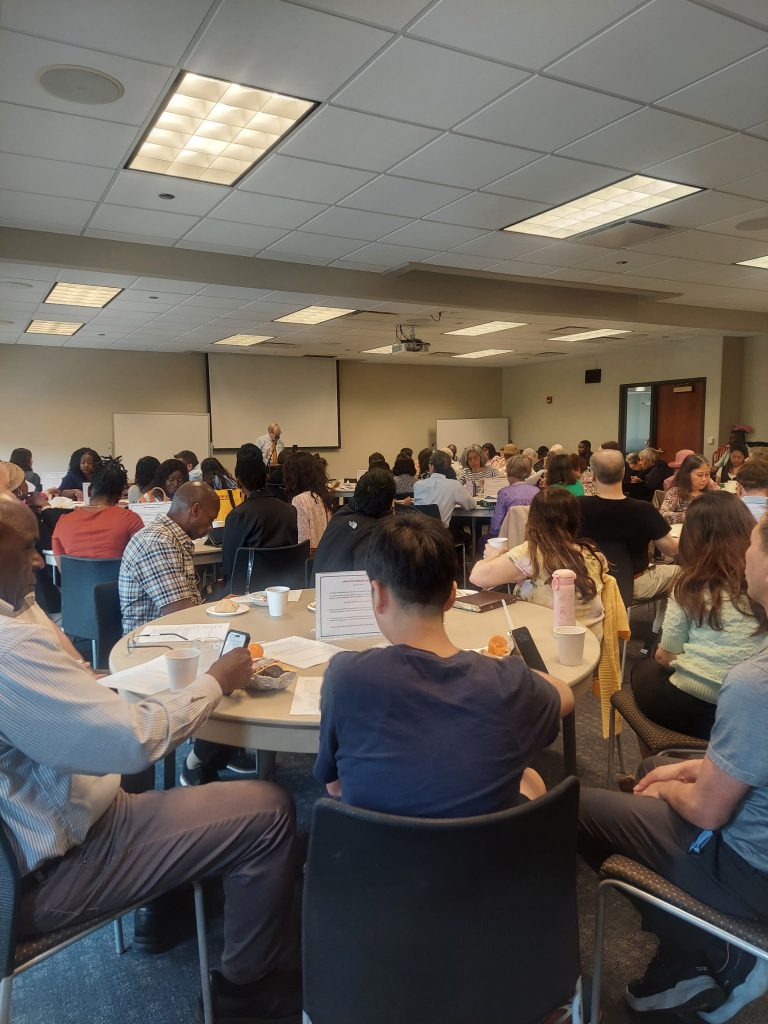 ATM Class1 Crowded conference room with attendees seated at round tables, eating and taking notes as a speaker stands at the front.