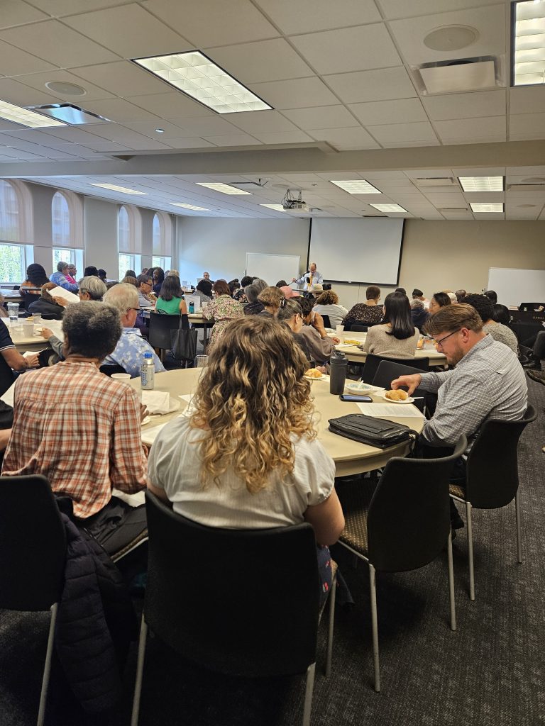 ATM Class2 A large meeting room full of people seated around round tables, a speaker at the front near a projector screen and whiteboard giving a talk.