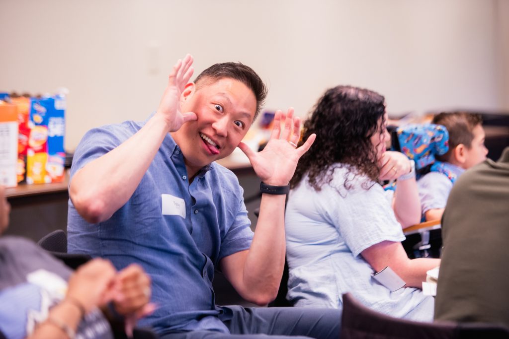 Toby Man in a blue shirt playfully frames his face with hands in a classroom setting, smiling at the camera.