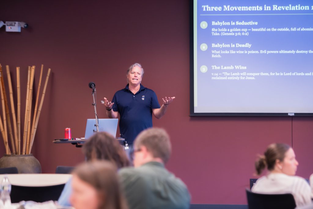 Mark Teaching #2 Speaker at a podium with a microphone gesturing as a slide titled 'Three Movements in Revelation' is projected on a blue screen to the right.