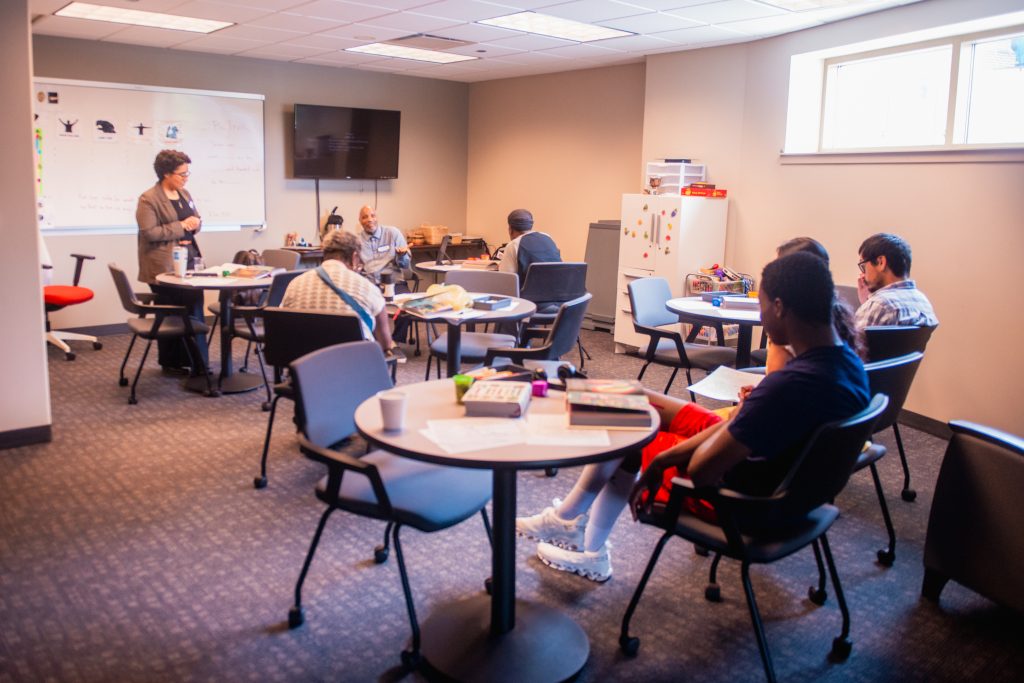 Co-Laborers #2 Group of adults seated around round tables in a classroom, a woman stands at the whiteboard at front.