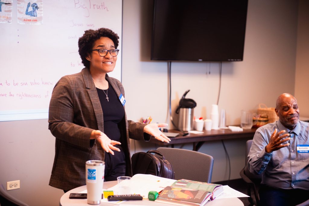 Liz & Antonio Teaching #1 Woman in a blazer presenting to a small group in a conference room, gesturing with open hands as others listen. Whiteboard, cups, and books on the table.