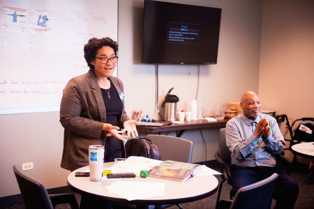 Liz & Antonio Teaching #2 Woman in blazer speaking to a small group in a conference room, with a whiteboard and TV screen behind her.